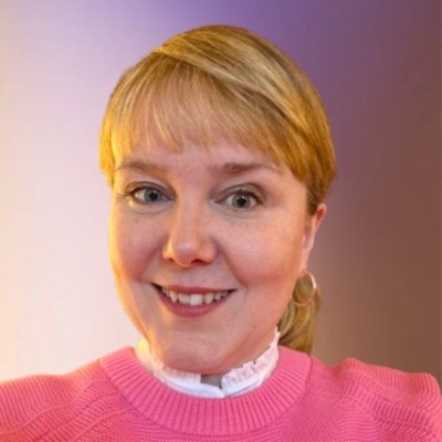 Photo of Angharad. She is a white woman, middle aged, with long fair hair. She's wearing a pink jumper and smiling at the camera.